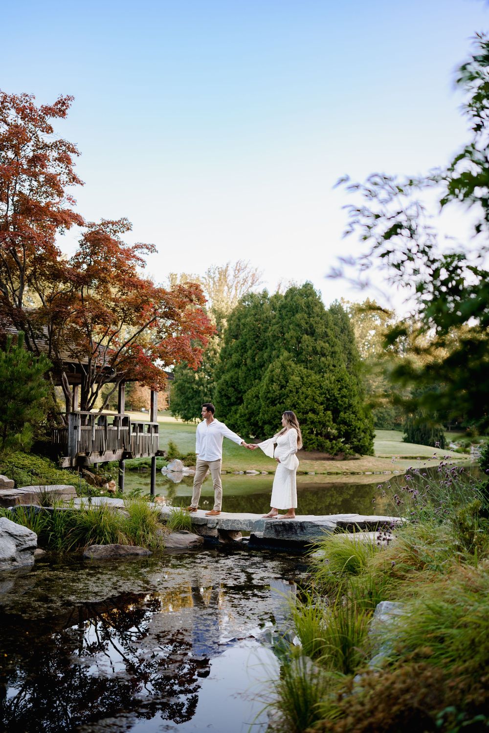 Couple holding hands on stepping stones over a serene garden pond surrounded by lush trees and greenery, during maternity session in MD