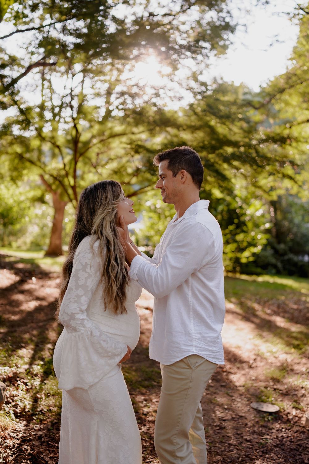 Couple in white outfits share a tender moment on a sunlit forest path during an outdoor maternity session in Wheaton, MD