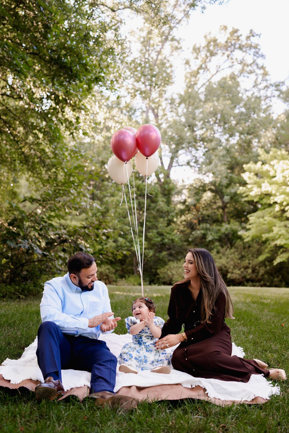 Family of three enjoying a picnic on grass with pink and white balloons in a lush green park, in Brookside Gardens, MD
