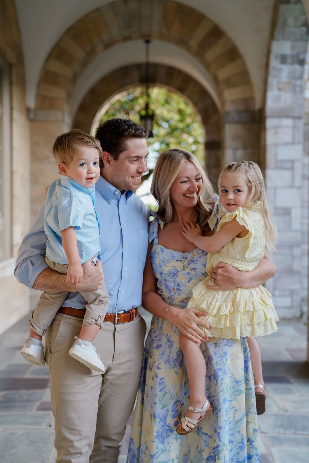 Happy family of four posing under stone archway, parents holding young children dressed in pastel spring outfits, in Ellicott City, MD