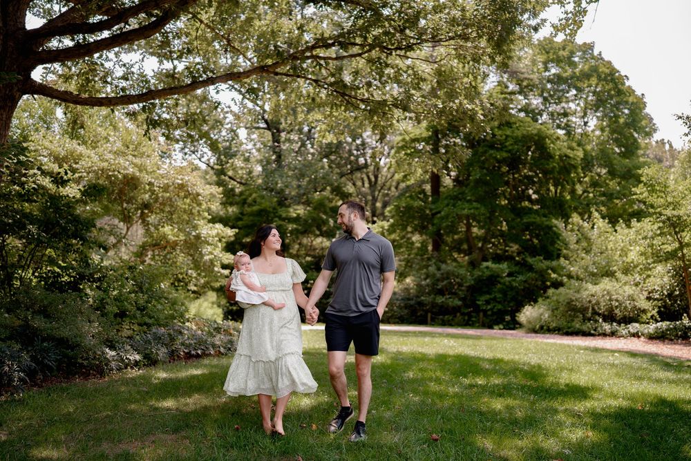 A couple holds hands while walking in a lush green park, with the woman in a white dress cradling a newborn baby, in Rockville, MD