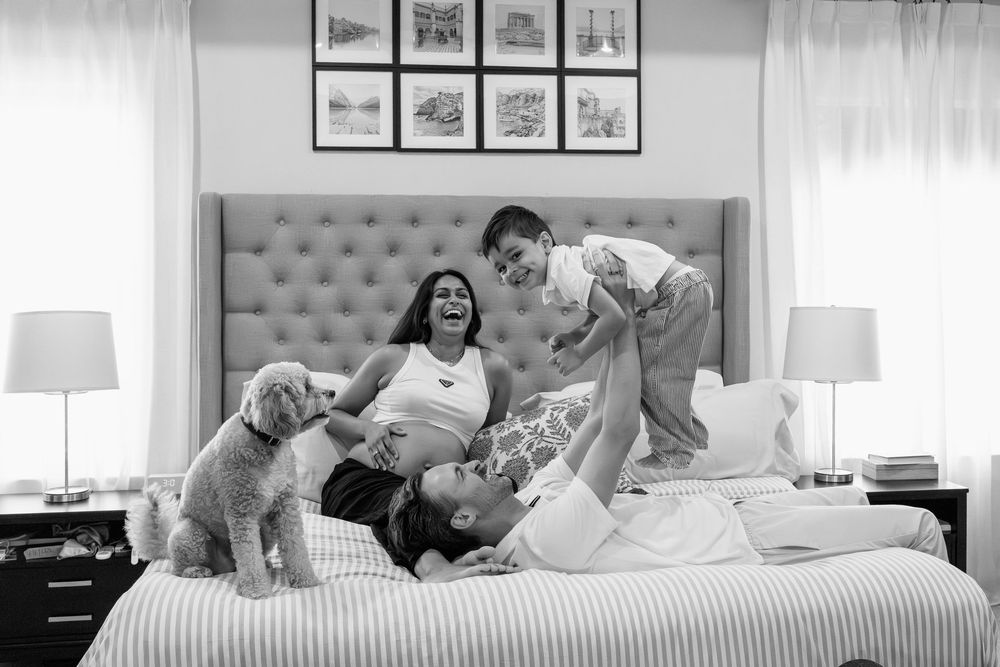 Black and white family photo of pregnant woman, man, child, and dog playing joyfully on a bed at home, in Odenton, MD