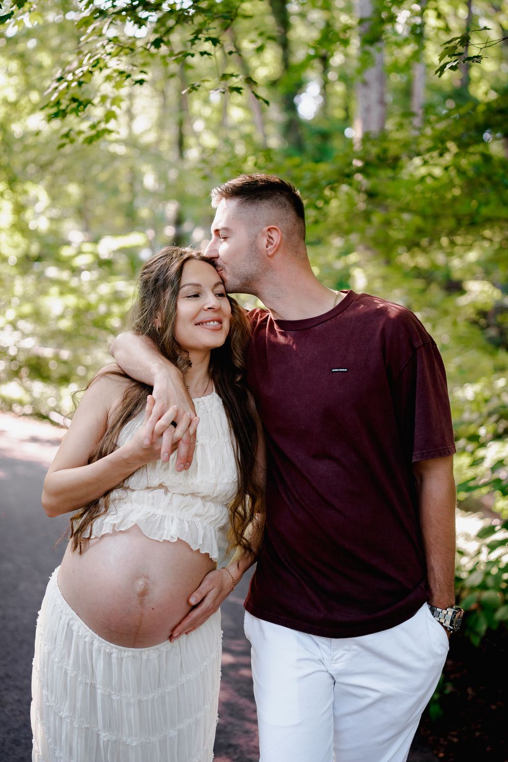 Pregnant woman in white dress with partner kissing her head outdoors in a lush green park setting, in Brookside Gardens, Maryland