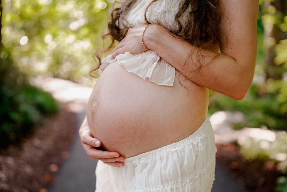 Pregnant woman in white dress cradling bare baby bump outdoors on a tree-lined path, in Maryland