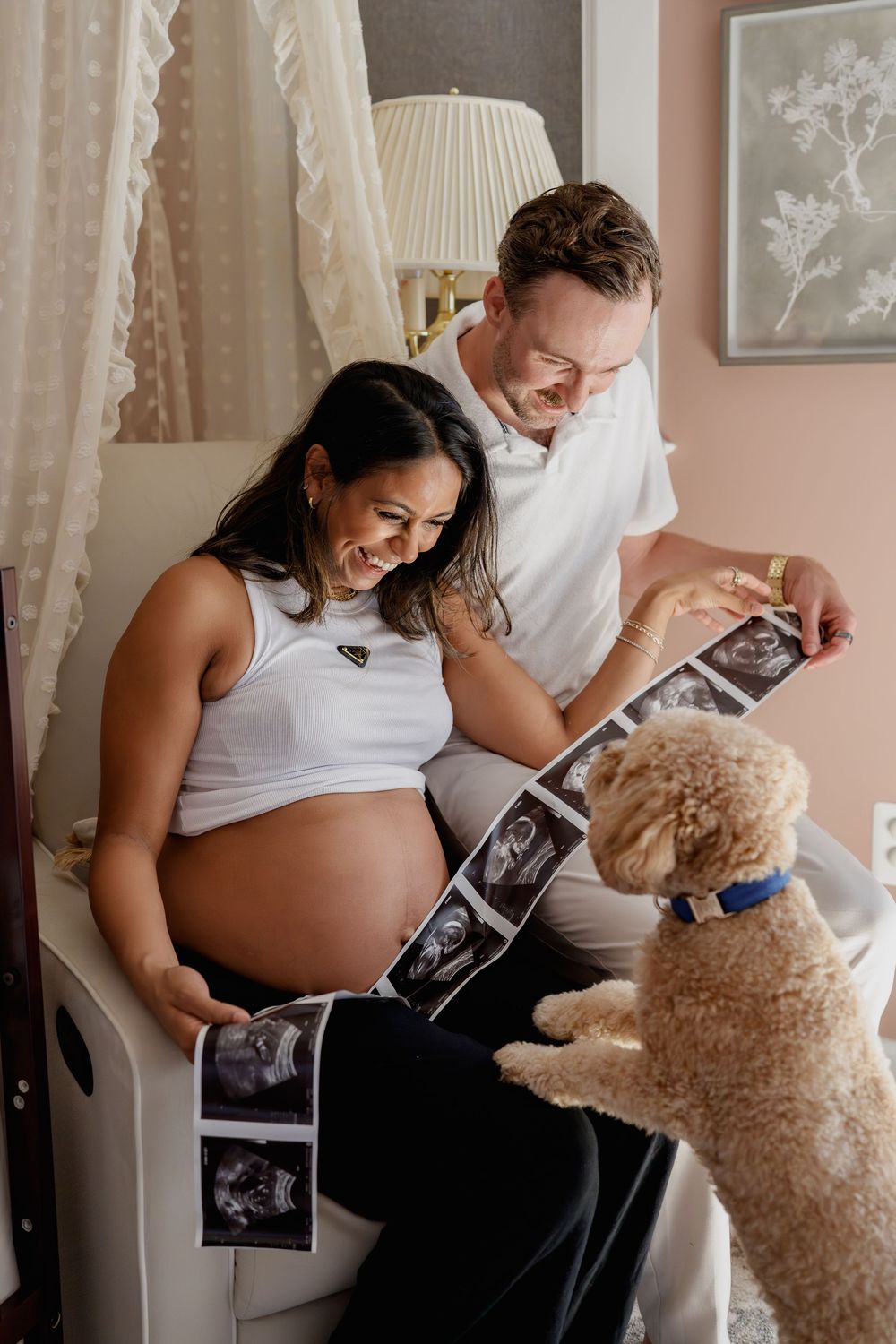 Pregnant woman and man smile at ultrasound photos while their curly golden dog looks on in a cozy nursery, in Odenton, MD
