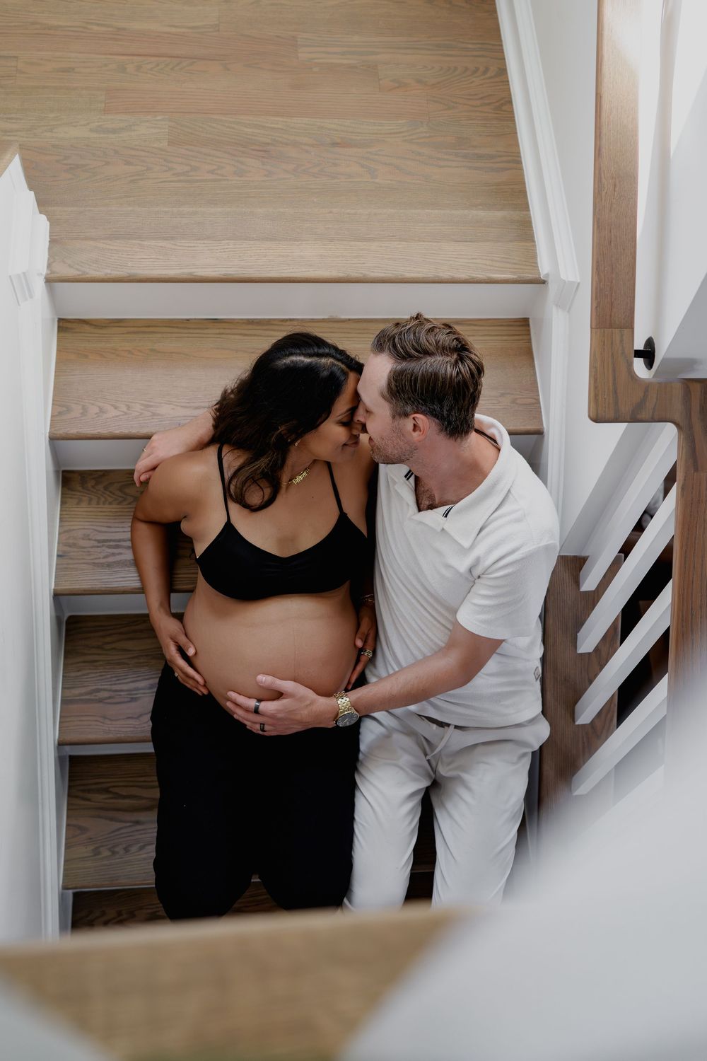 Couple sharing a tender kiss on wooden stairs, man gently holding pregnant woman's baby bump in a bright home, in Odenton, MD