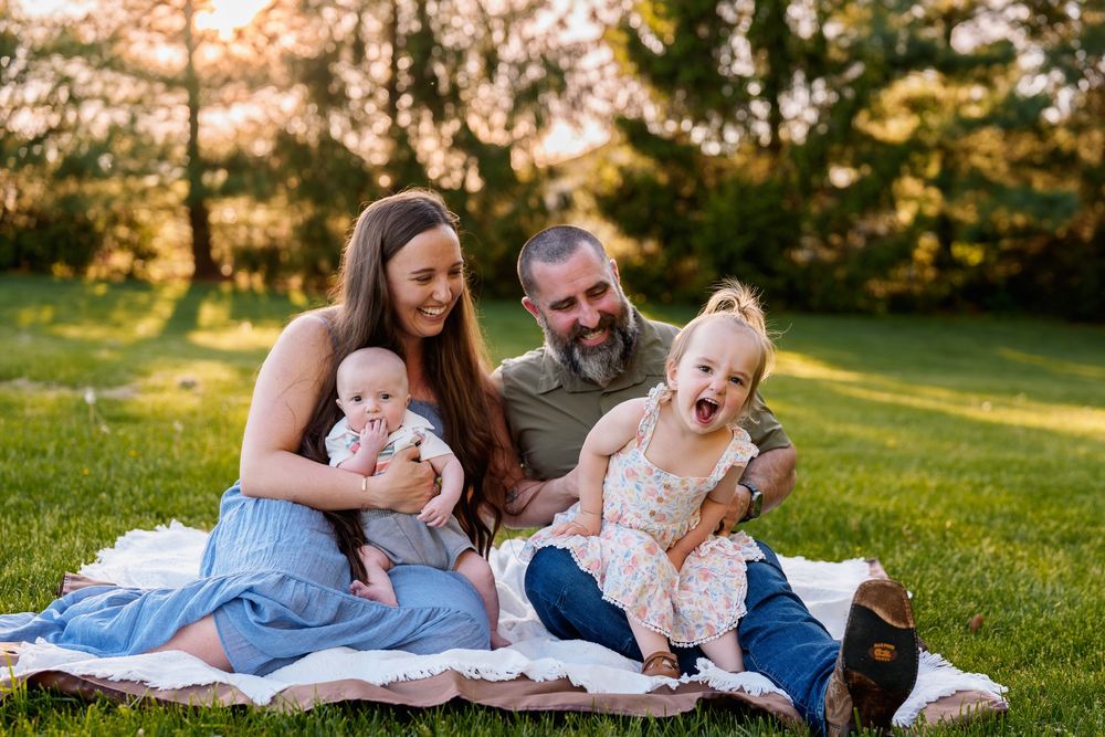 Happy family of four sitting on a blanket in a sunny green park, parents smiling with baby and laughing toddler girl, in Frederick, MD