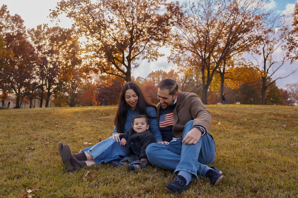 Family of three sitting on grass in autumn park, parents smiling at toddler holding small American flag, at Constitution Gardens in DC