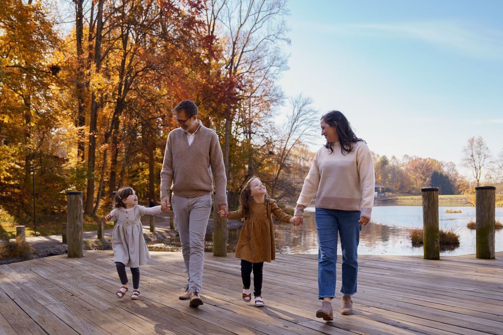 Family of four walking hand-in-hand on a wooden dock surrounded by colorful autumn trees near a lake, in Columbia, MD