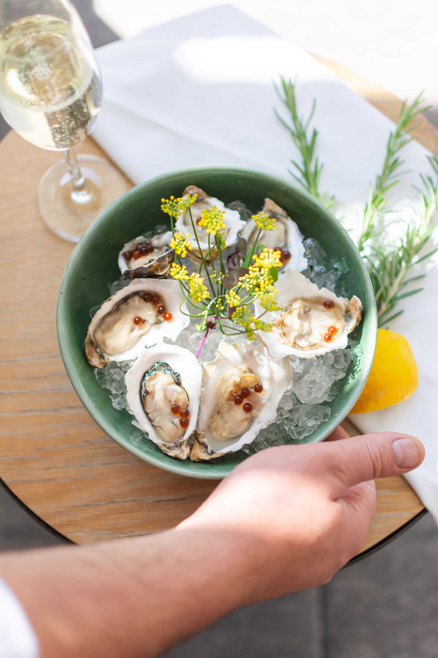 Fresh oysters on ice garnished with yellow flowers and herbs served in a green ceramic bowl with lemon wedge.