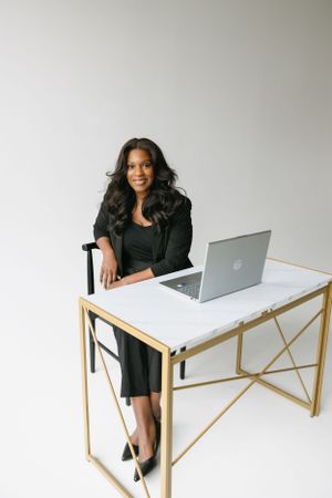 Professional woman sits at modern white and gold desk with laptop in minimalist office setting.