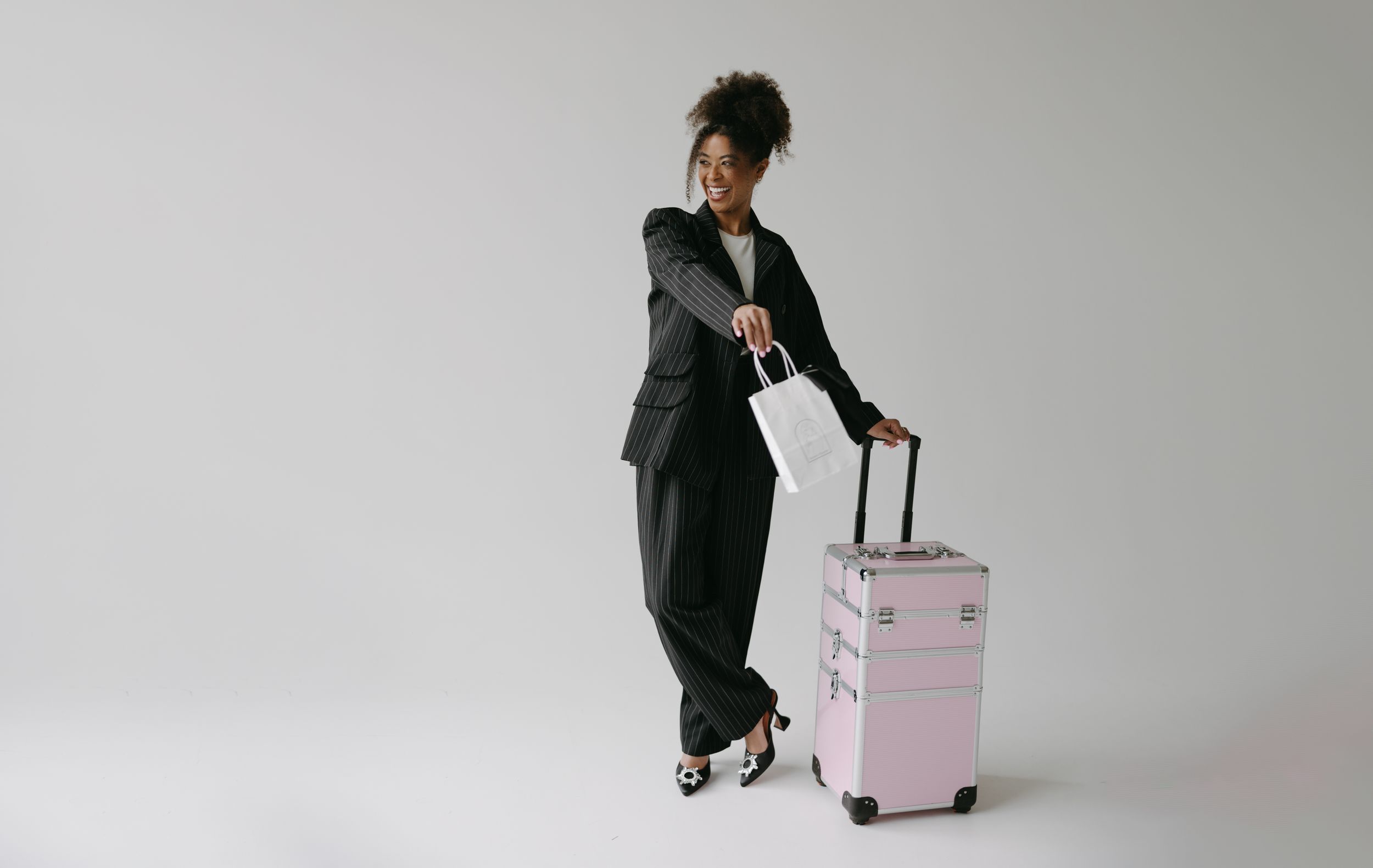 Stylish woman in business attire posing with pink rolling luggage against white background.