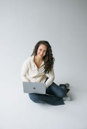 Person in white sweater works casually on laptop while sitting on floor against neutral background.