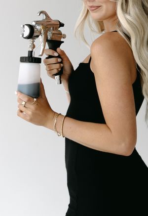 Woman in black dress holding a paint sprayer equipment against white background.