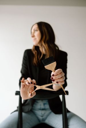 A hairstylist dressed in black holds professional scissors and a comb against a light background.