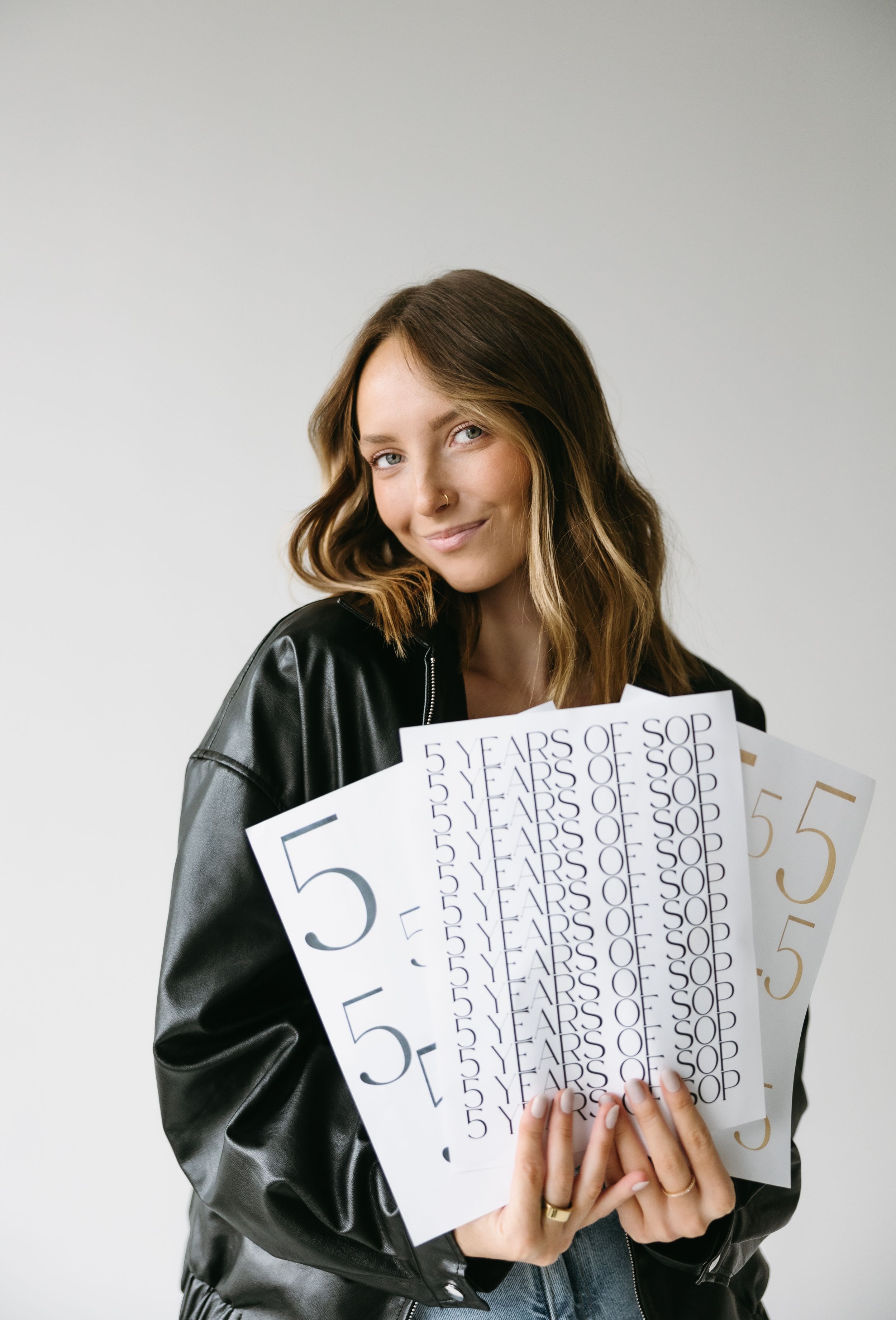Woman in leather jacket holds number designs and typography samples against white background.