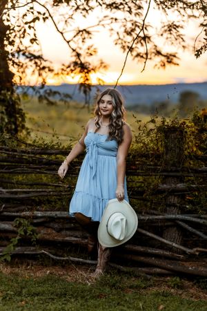 A person in a light blue dress poses against rustic wooden fencing during golden hour sunset.