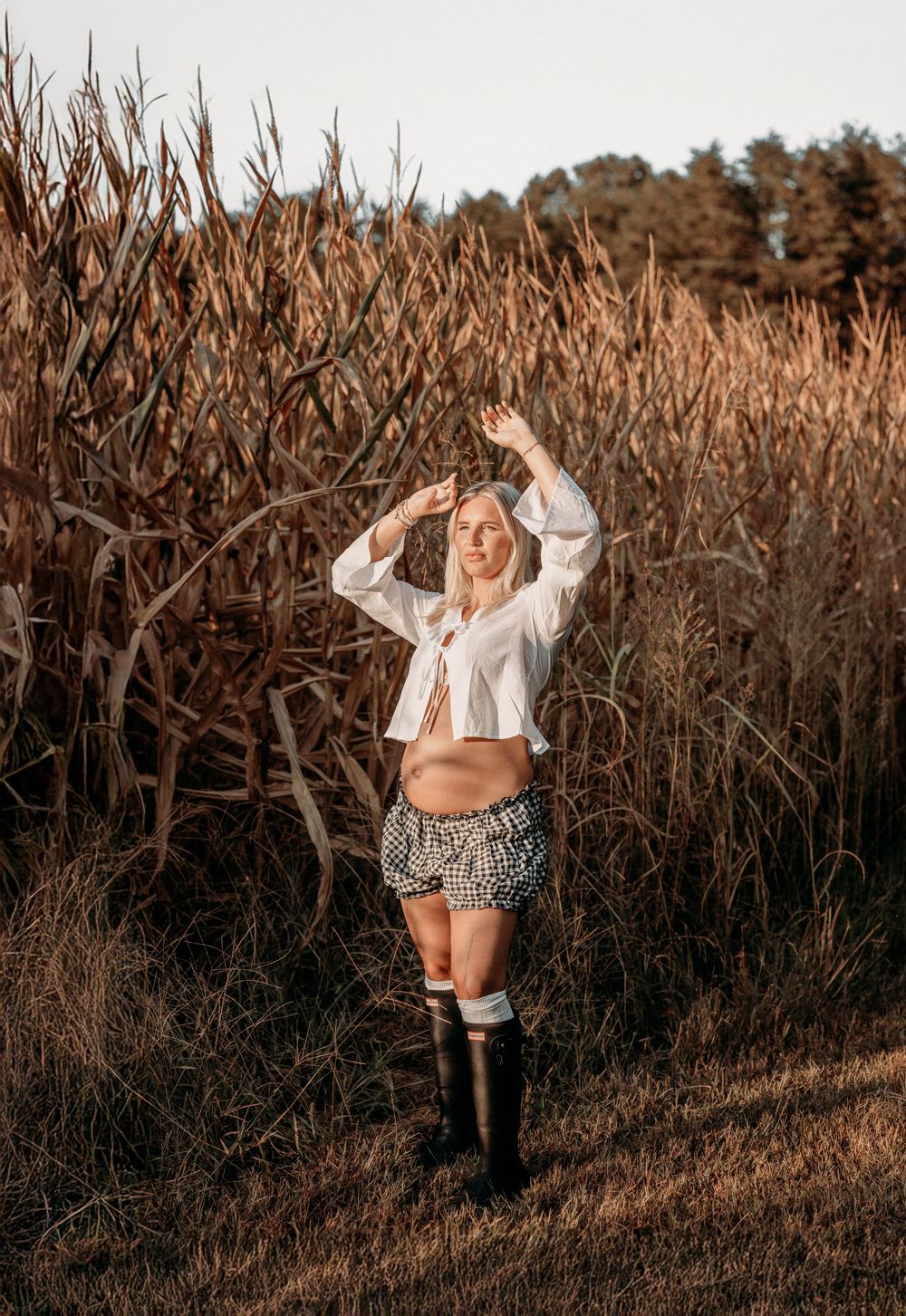 A figure poses in a white crop top and patterned shorts against a backdrop of tall dried corn stalks at sunset.