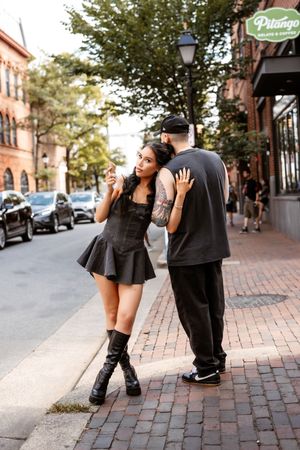 A romantic couple shares playful moments on a brick sidewalk in an urban setting with storefronts and trees.