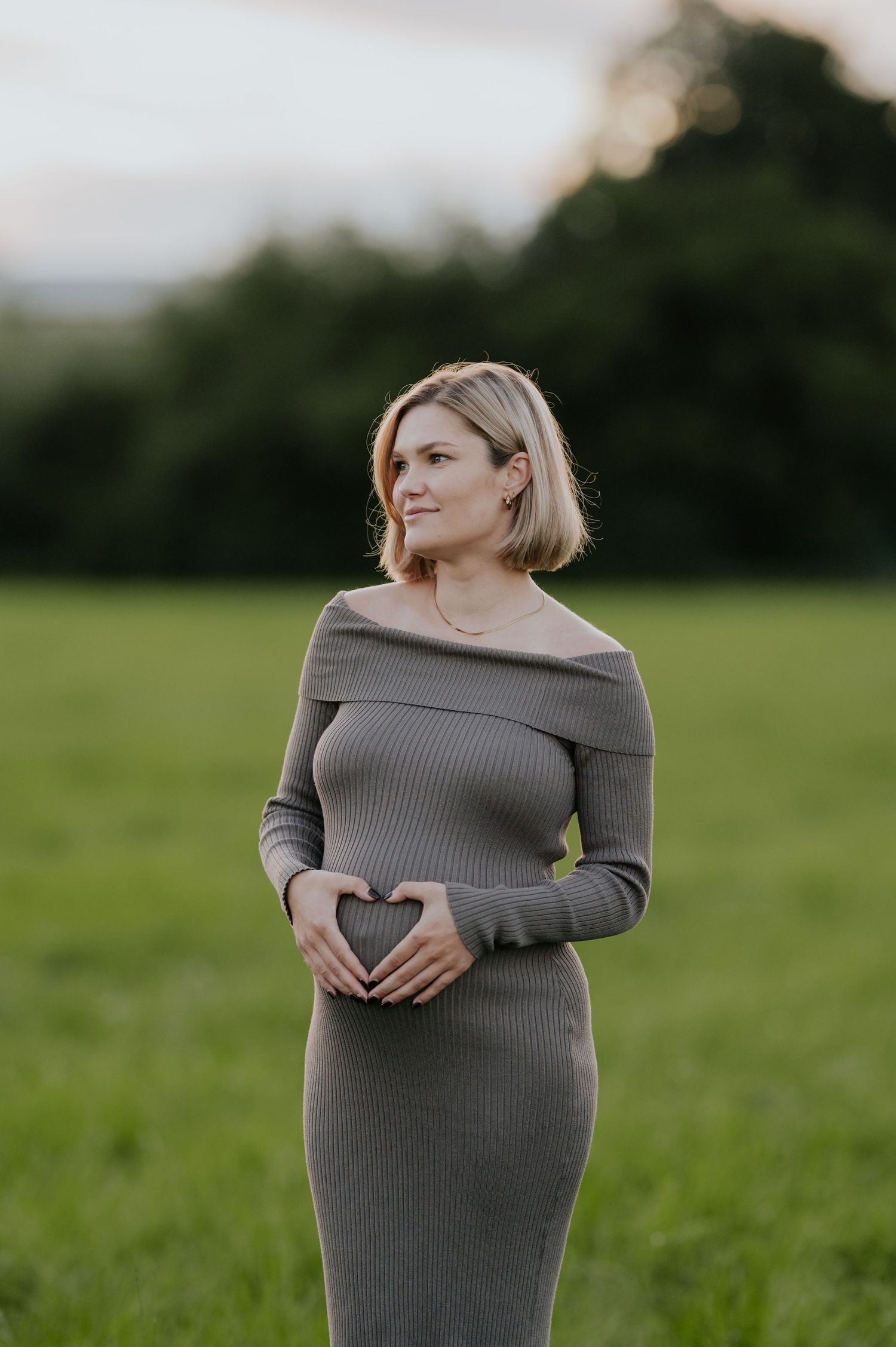 Eine schwangere Frau in einem grauen Strickkleid steht während eines Schwangerschaftsfotoshootings auf einer grünen Wiese.