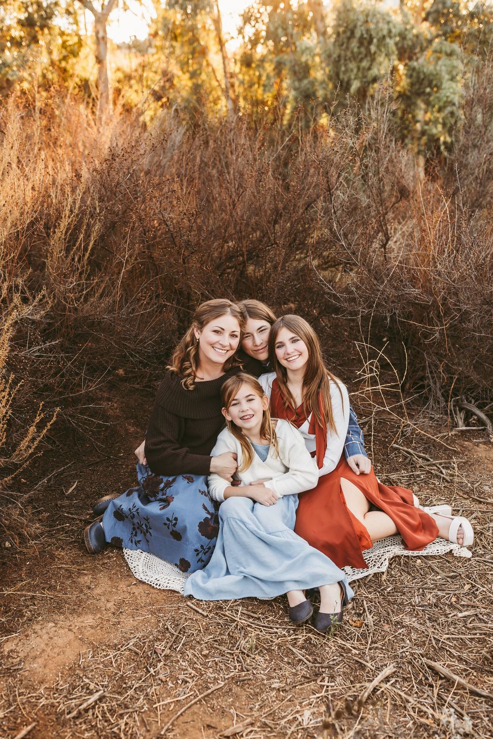 Three people relax together on autumn ground wearing coordinating dresses in light blue and rust colors during sunset.