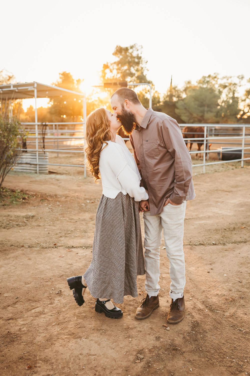A couple shares a romantic moment at sunset on a dusty ranch with wooden fencing in the background.