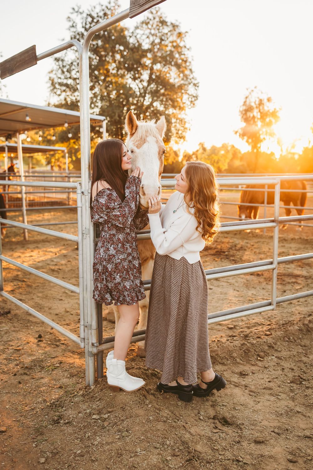 Two people pet a white horse in a fenced paddock at sunset while wearing dresses.