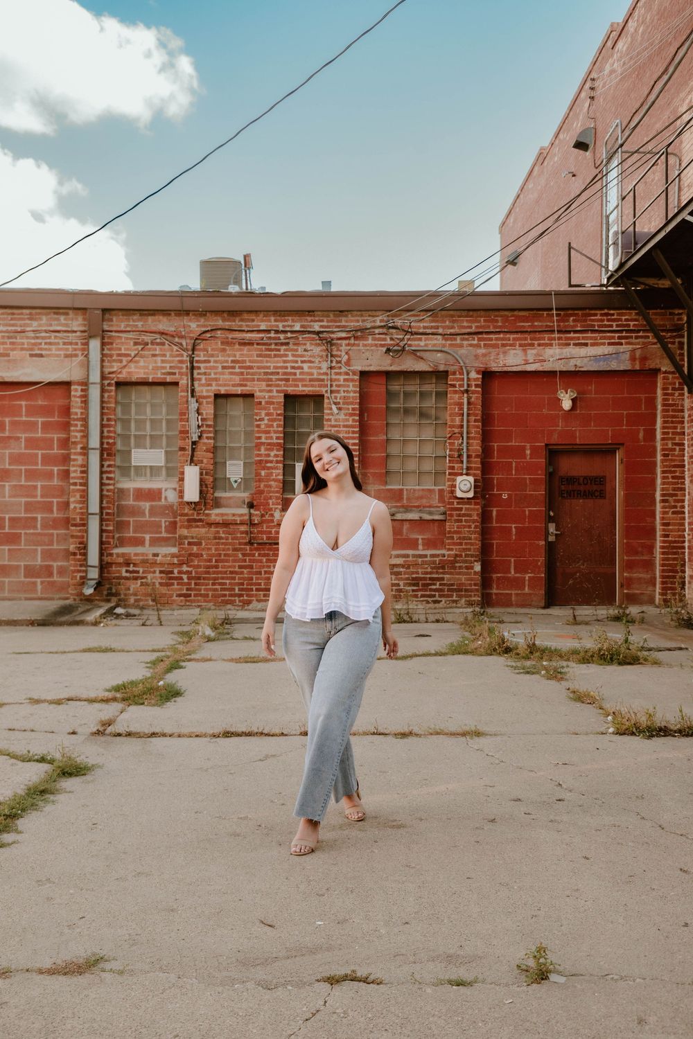 Young woman in white top and gray flared jeans posing confidently in an urban alley with red brick walls.