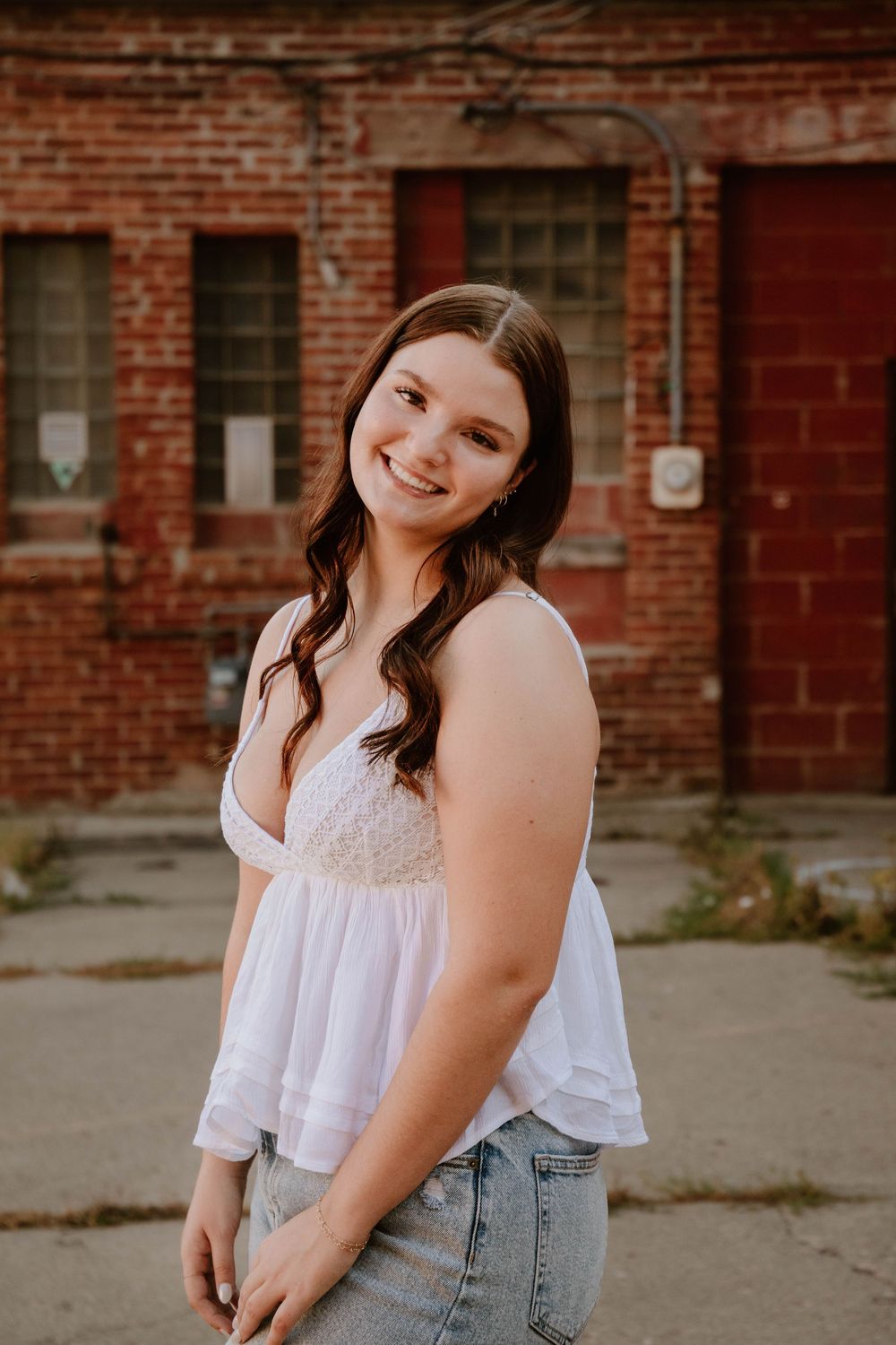 Smiling young woman in white top and jeans posing outdoors in front of a rustic red brick building.