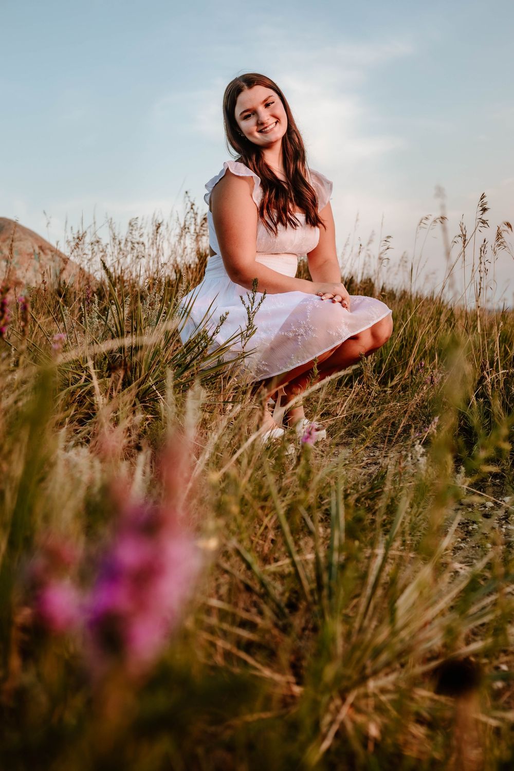 Young woman in white dress sitting in a wildflower meadow, smiling warmly in golden hour sunlight.