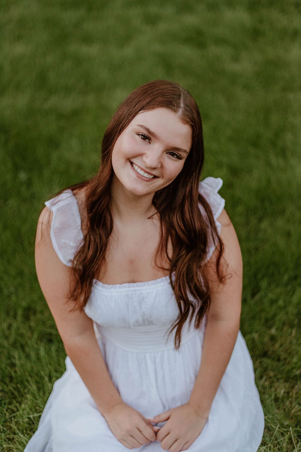Young woman with long auburn hair smiling in a white sundress, seated on green grass outdoors.