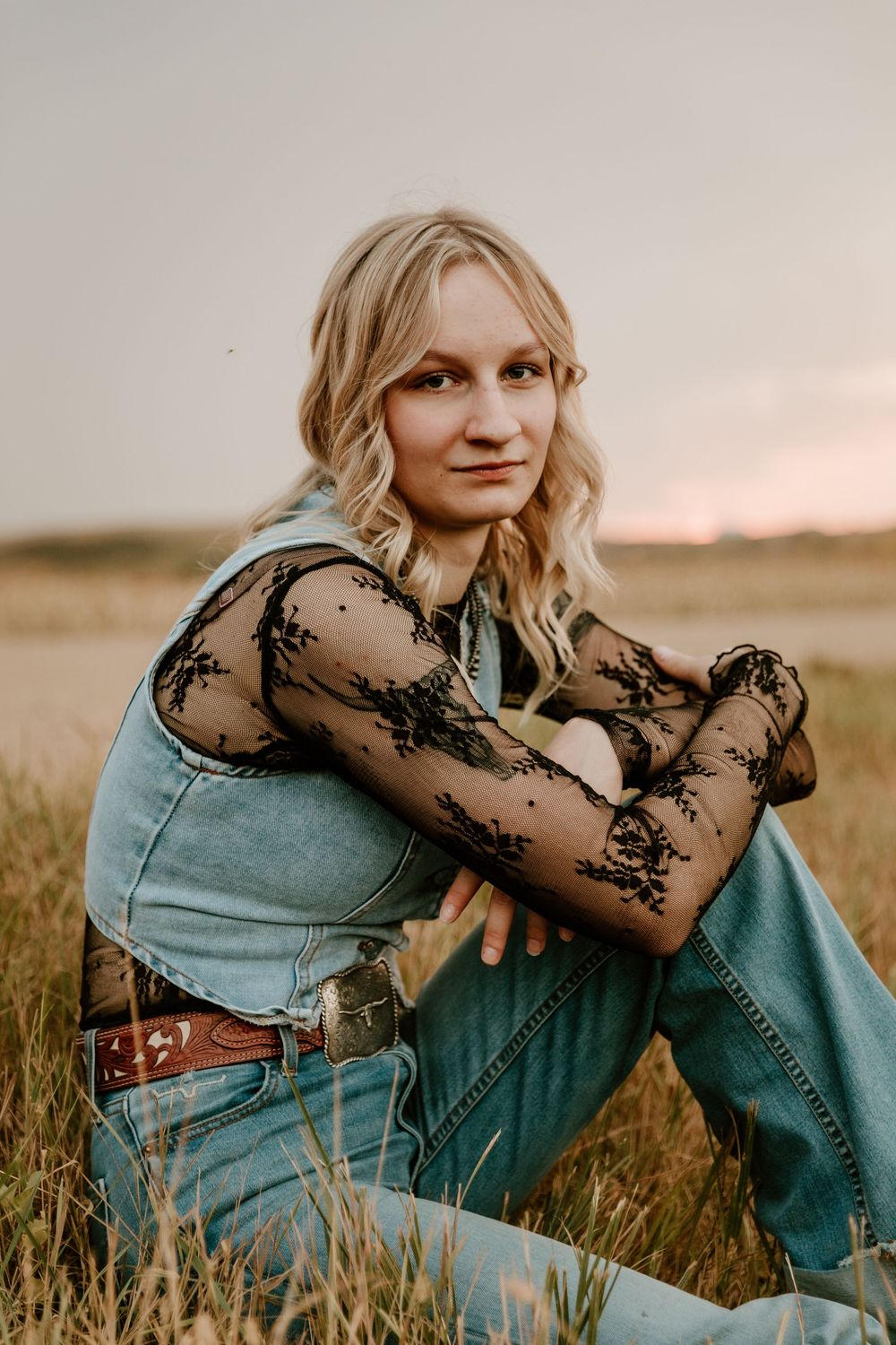 Blonde woman in denim overalls and black lace top sits in golden field at sunset, arms crossed on knees.