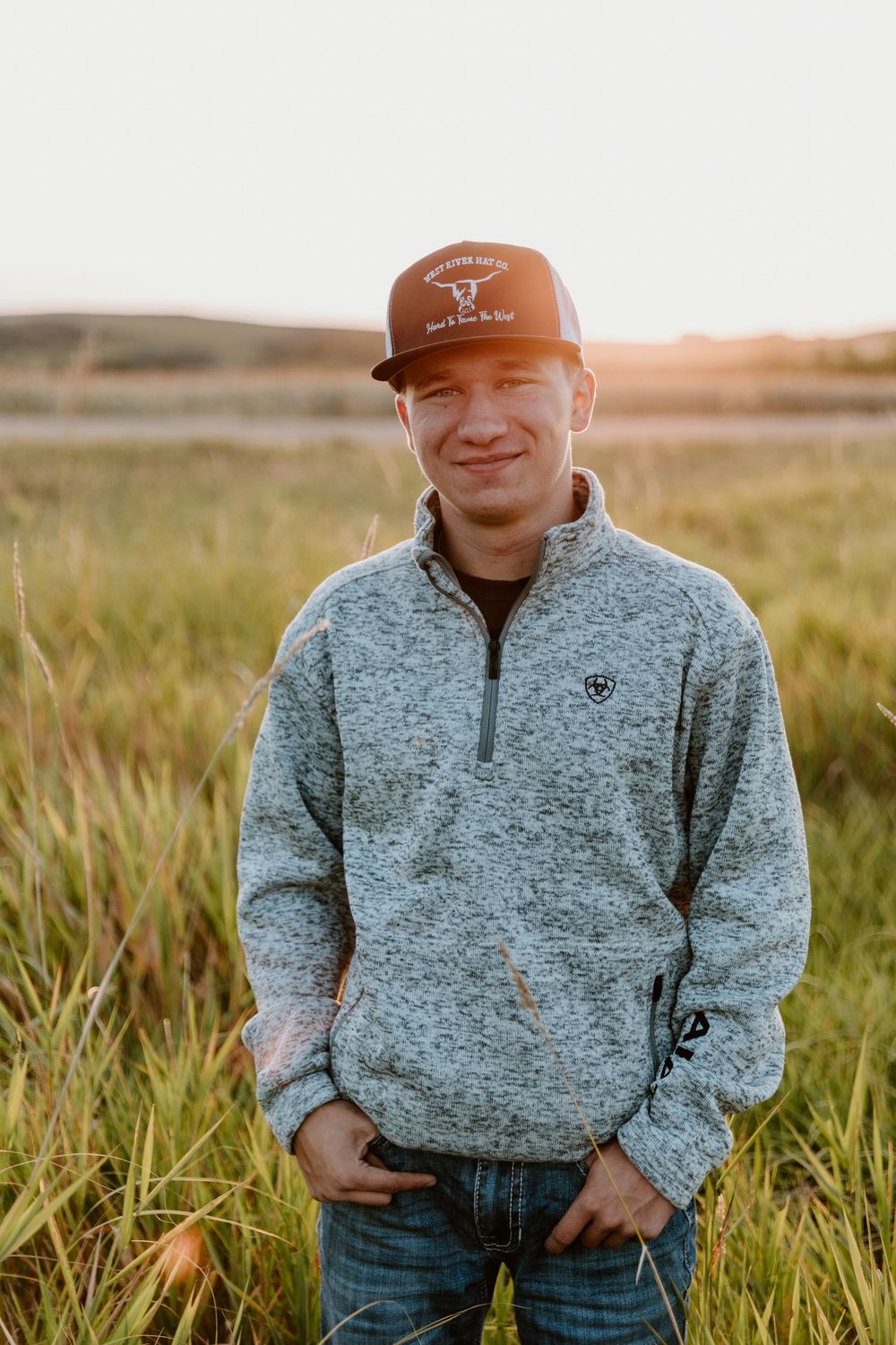 Person wearing a baseball cap and gray fleece pullover smiling in a grassy field at golden hour.
