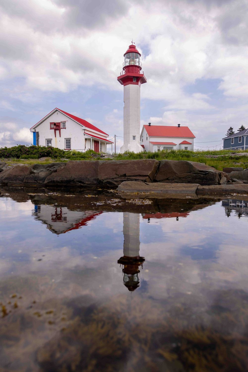 Red and white lighthouse with buildings reflected in tidal pool at Métis sur mer Site in Quebec.