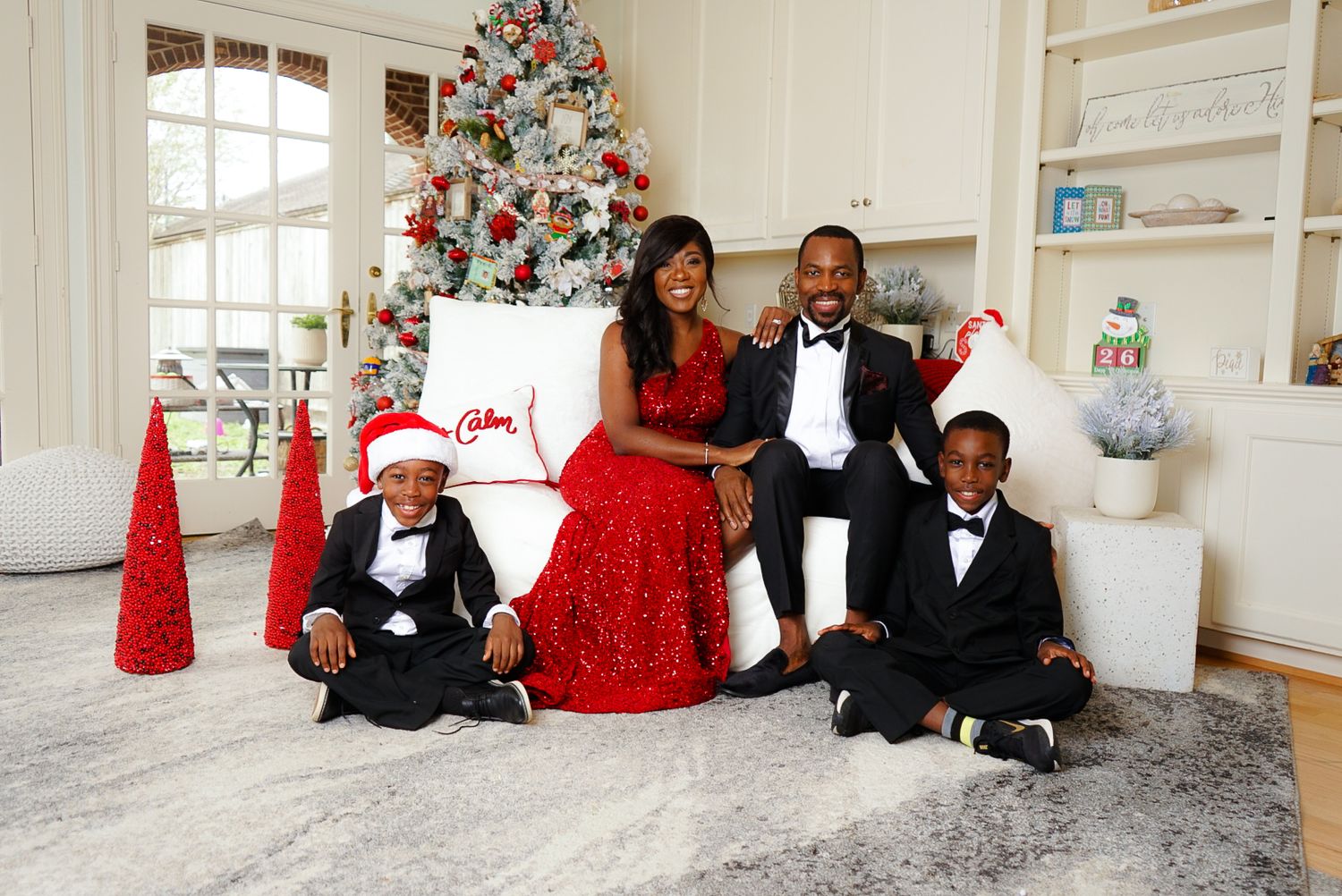 Family of four in formal attire poses by a decorated Christmas tree in a festive holiday portrait setting.