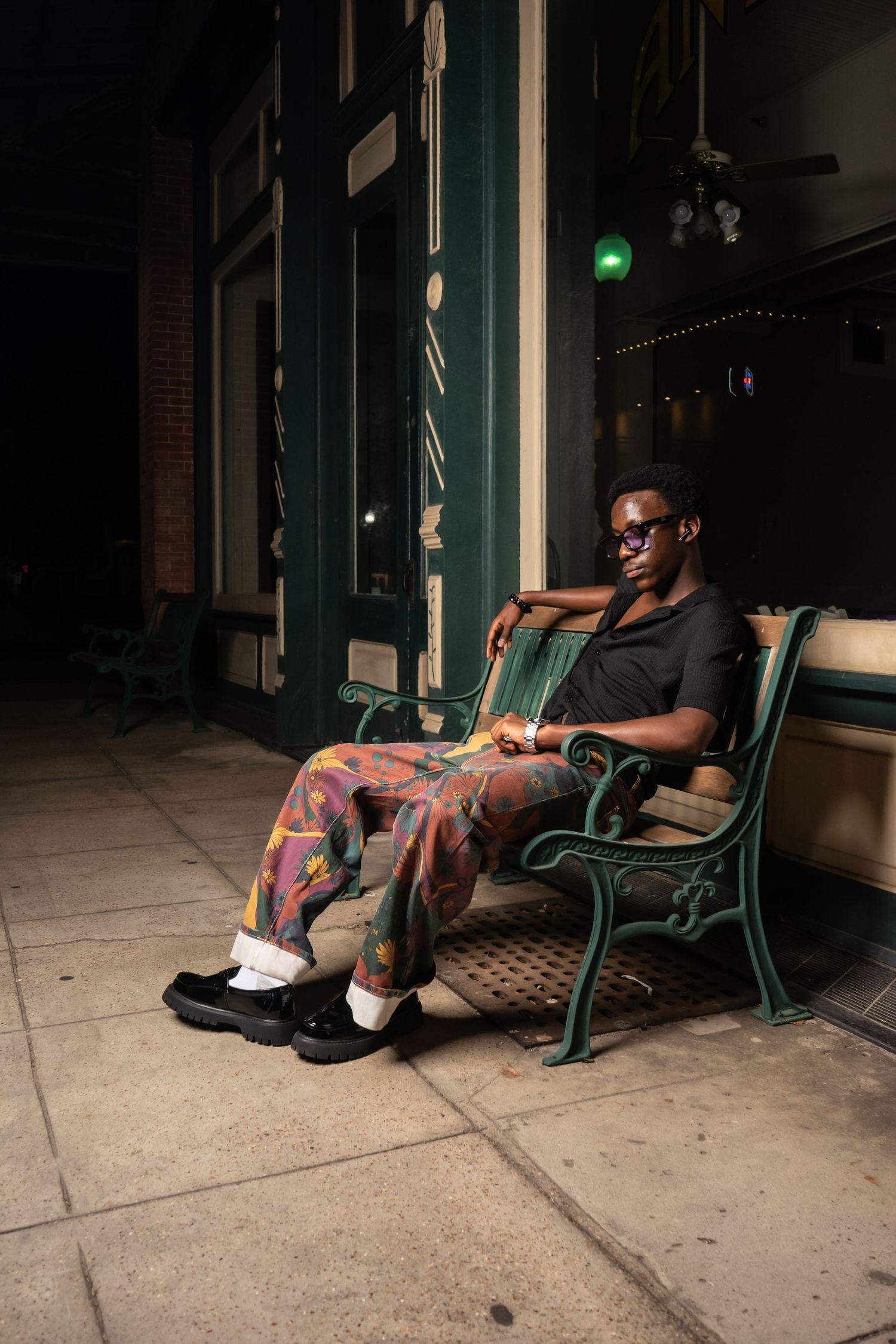 A stylish young Black man in colorful pants and sunglasses relaxes on a green iron bench outside a storefront at night.