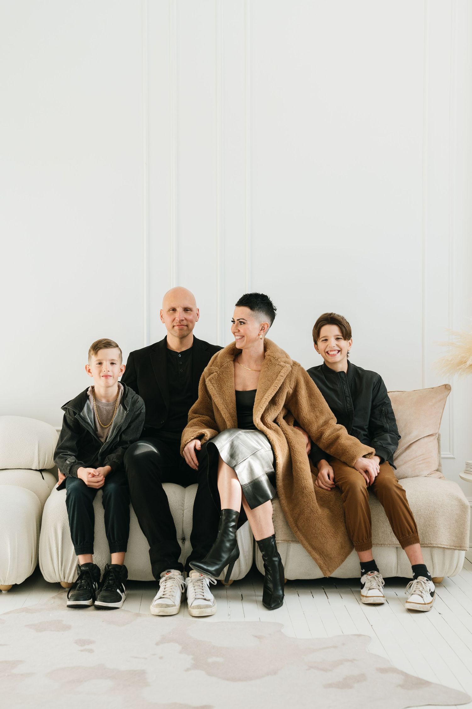 Four people sitting casually together on a beige couch in a minimalist room wearing stylish modern clothing.