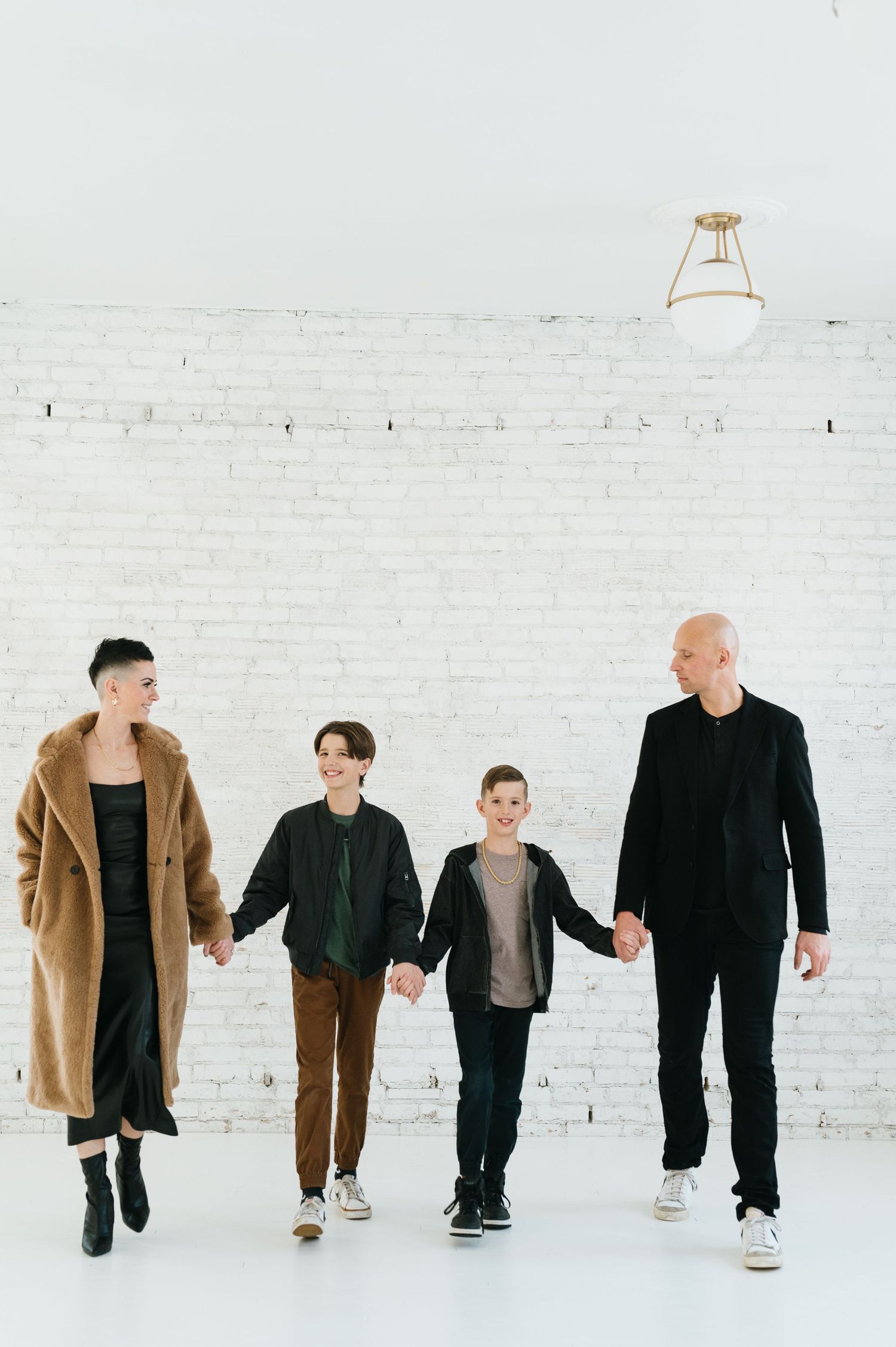 Four people holding hands while walking together against a white brick wall in a minimalist setting.