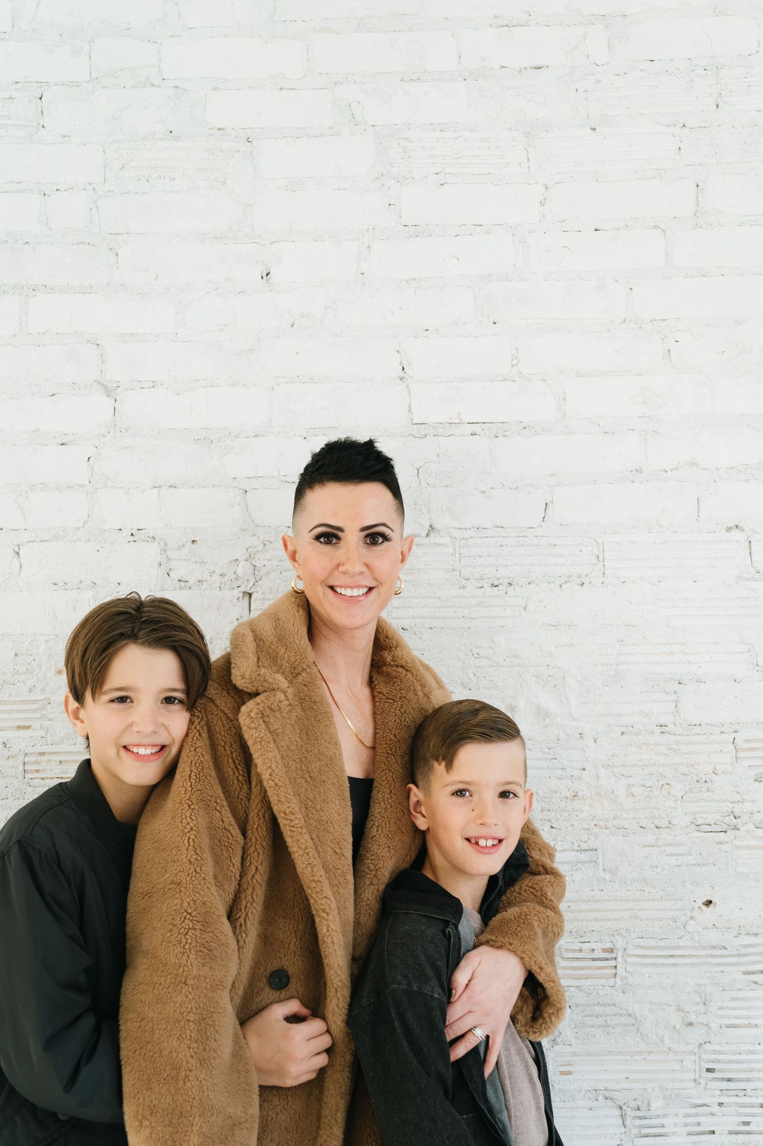 Family portrait against white brick wall with parent in brown coat and two children in dark sweaters.