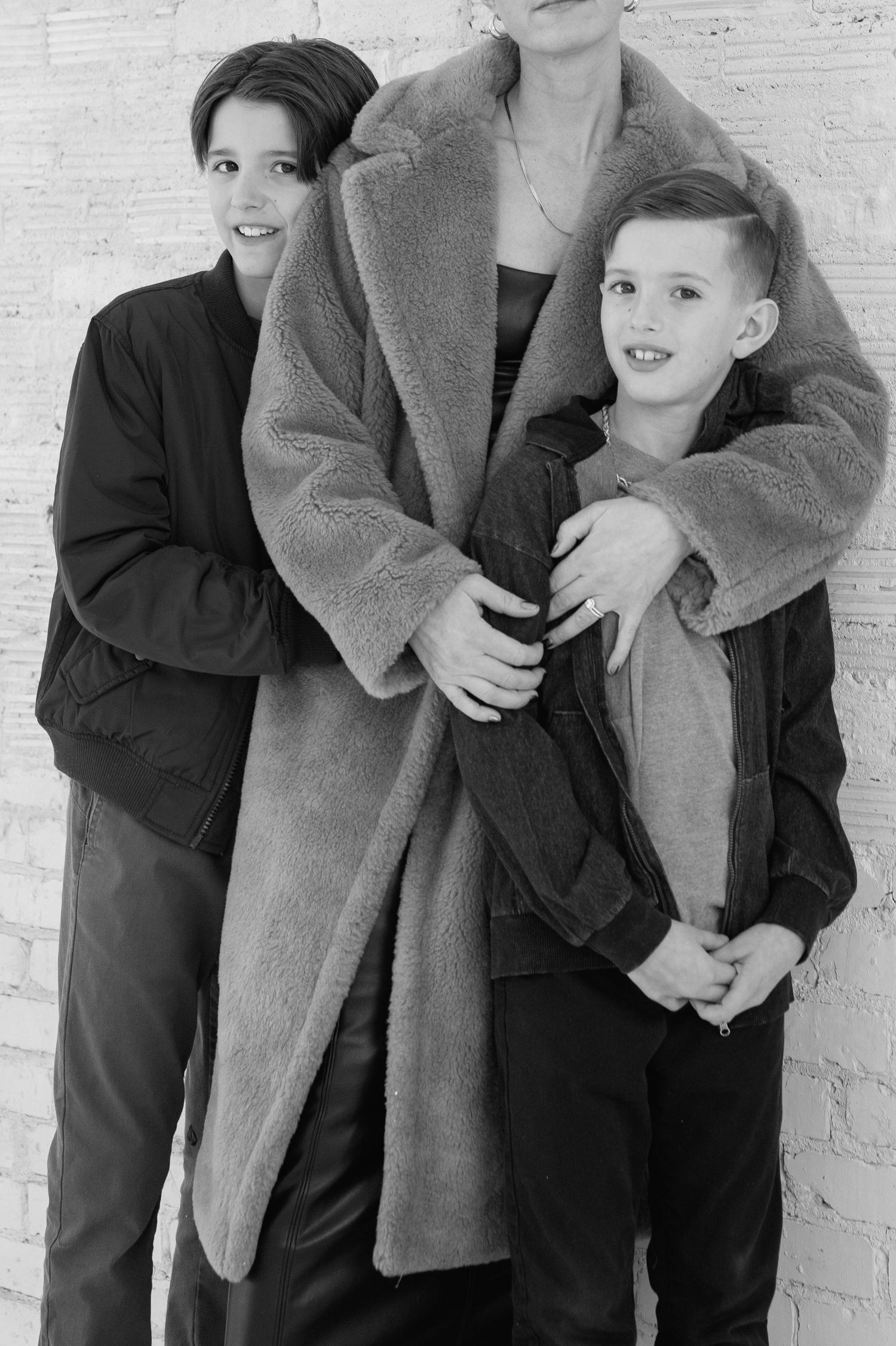A black and white portrait of three people wearing oversized coats against a light brick wall background.