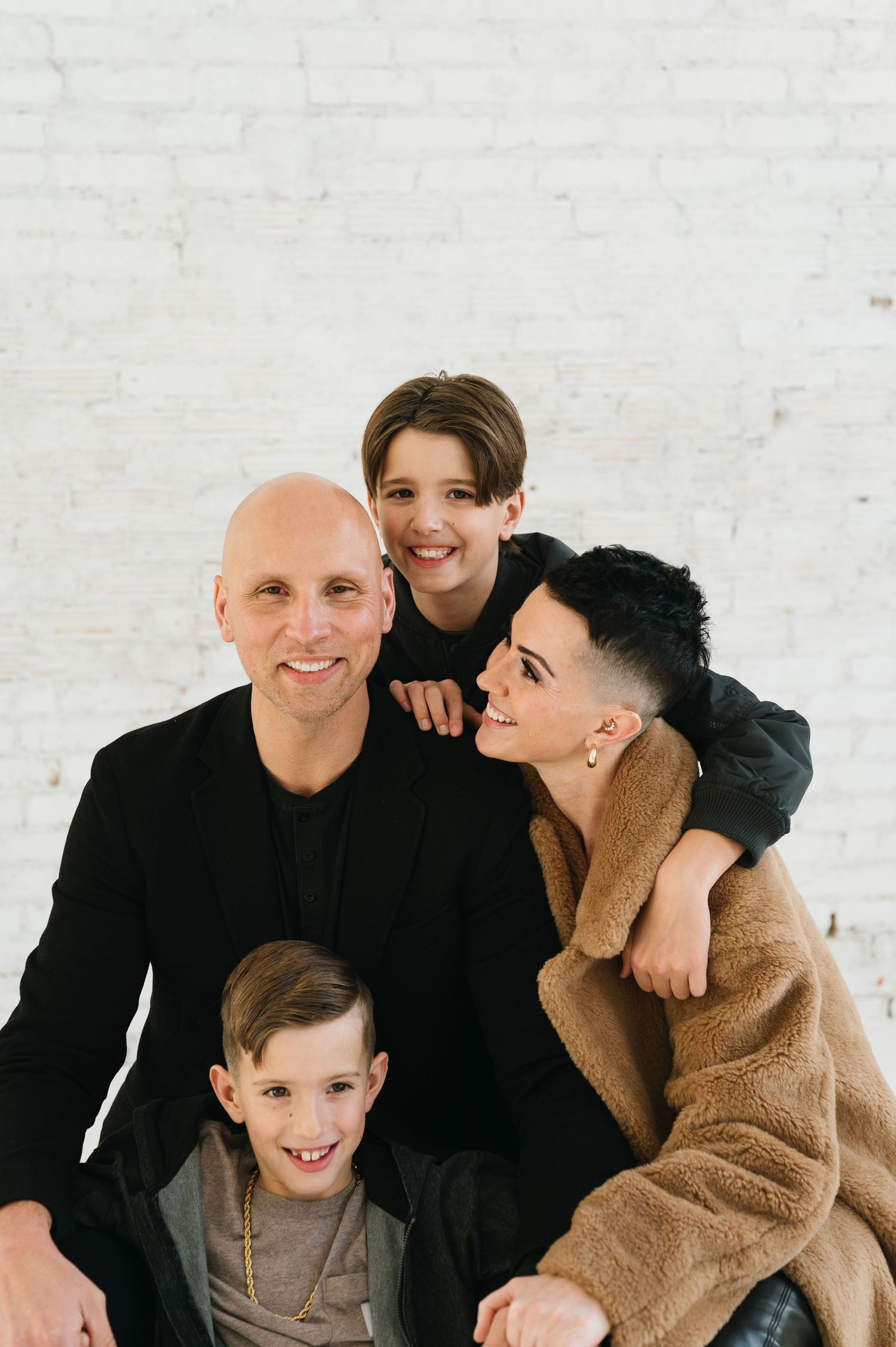 Family portrait with four people wearing casual attire against a white brick wall background.
