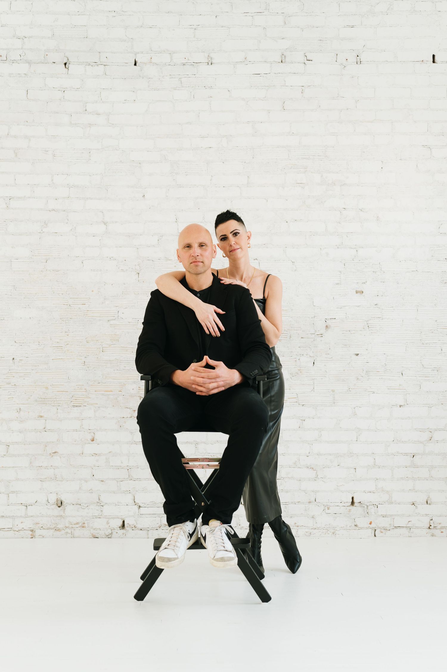 Two people dressed in black sit together on a stool against a white brick wall in a minimalist setting.