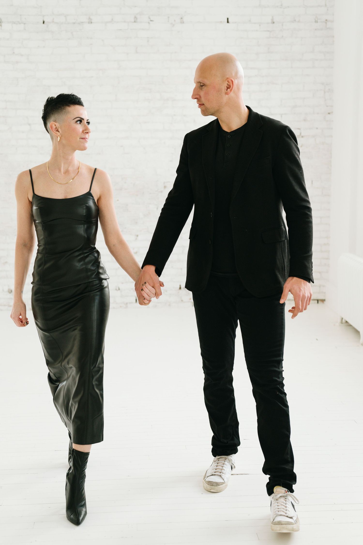A couple in coordinated black outfits holds hands while walking together in a minimalist white studio space.