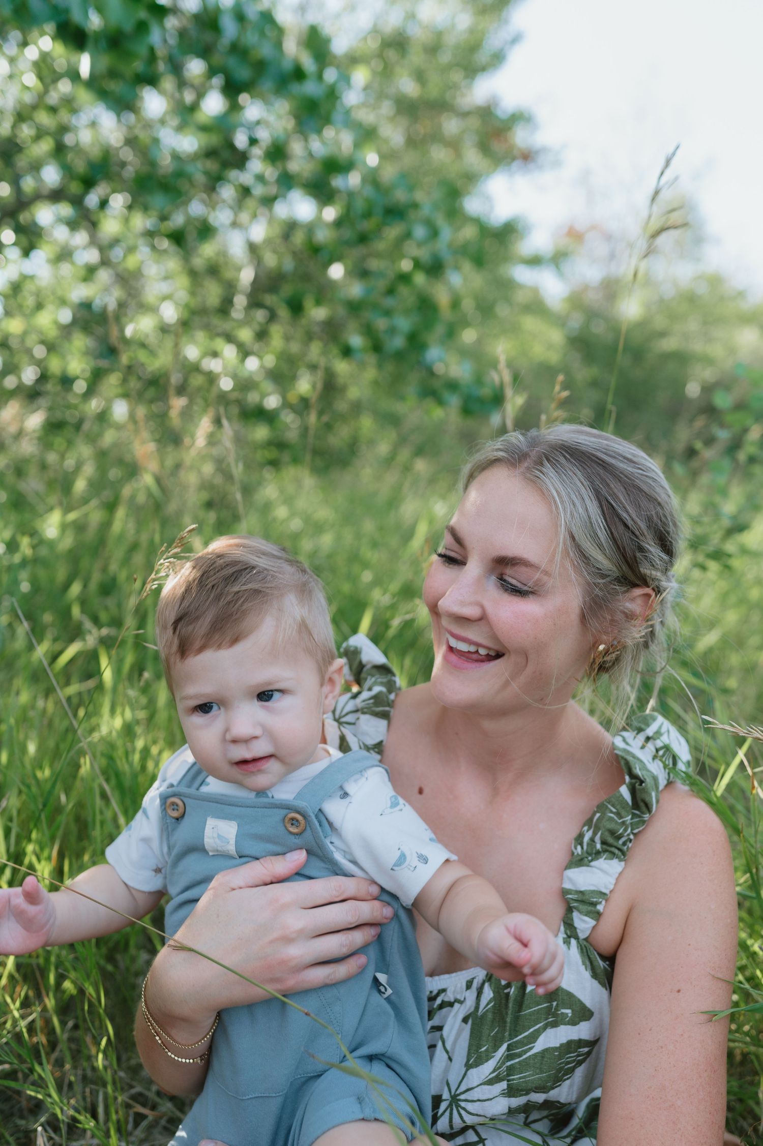 Summer portrait of parent and baby in overalls amid lush green foliage.
