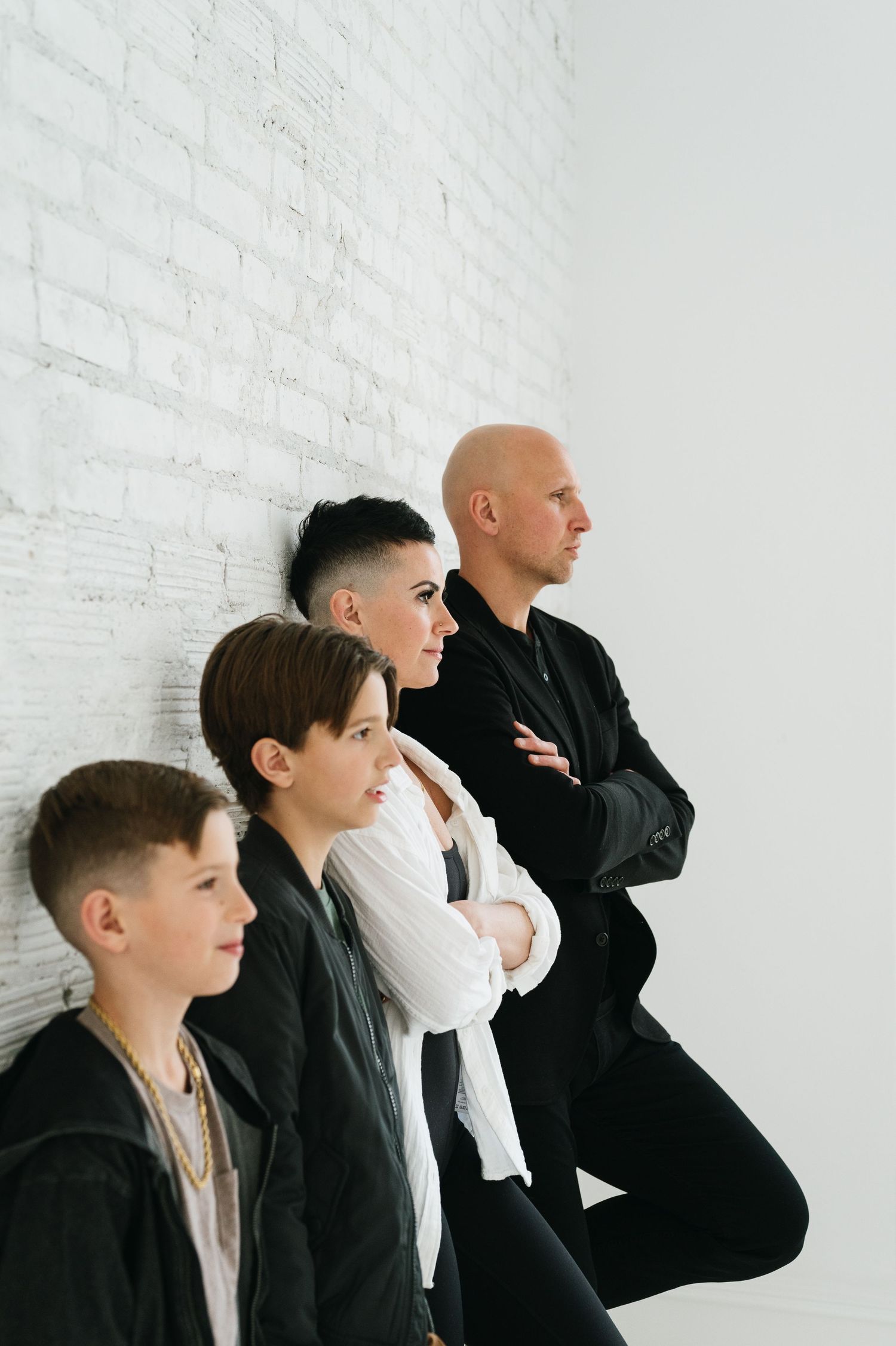 Four people posing in stylish black outfits against a white brick wall in an artistic profile shot.