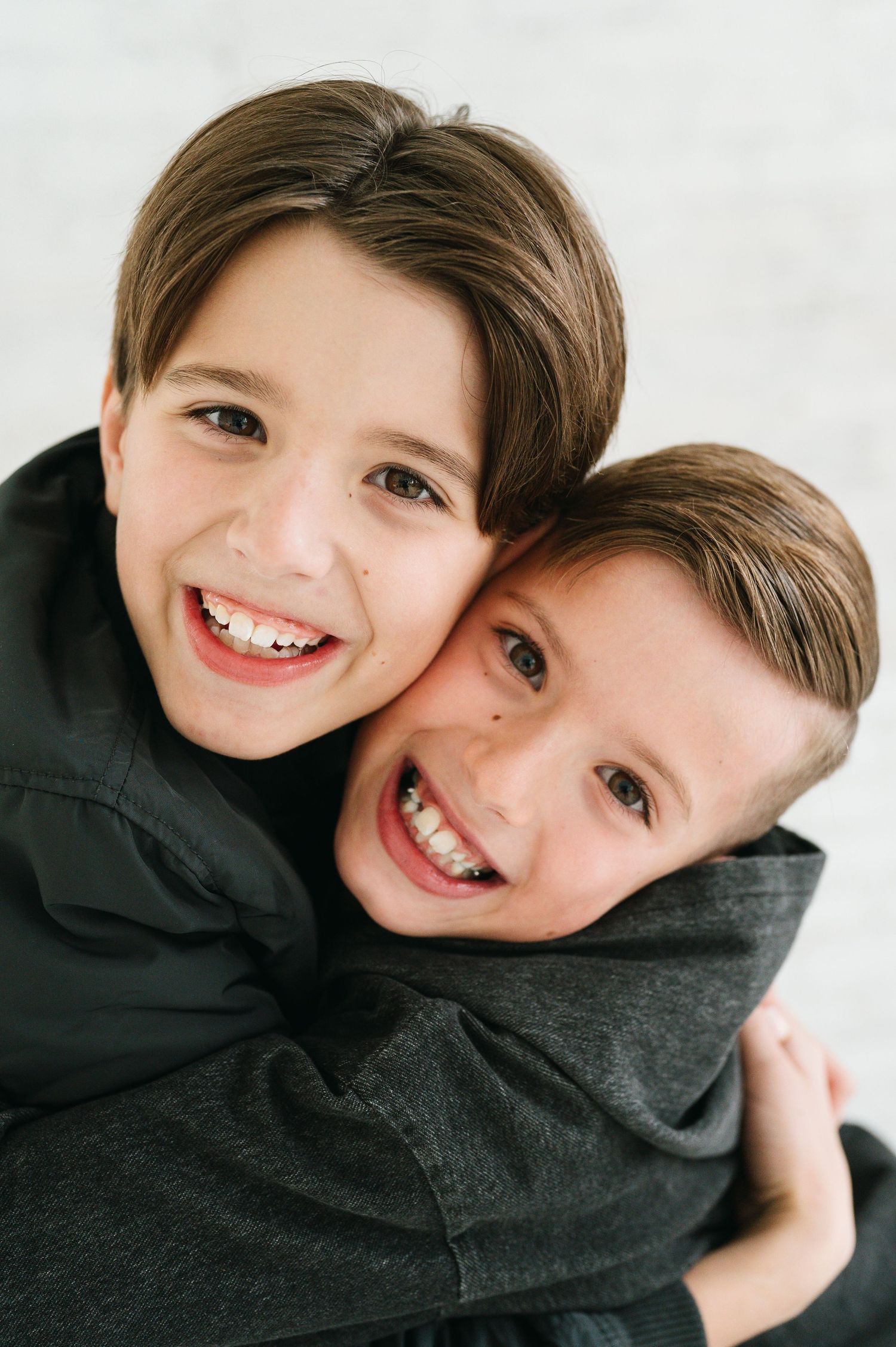 Two children wearing dark sweaters embrace while showing bright, cheerful smiles against a light background.
