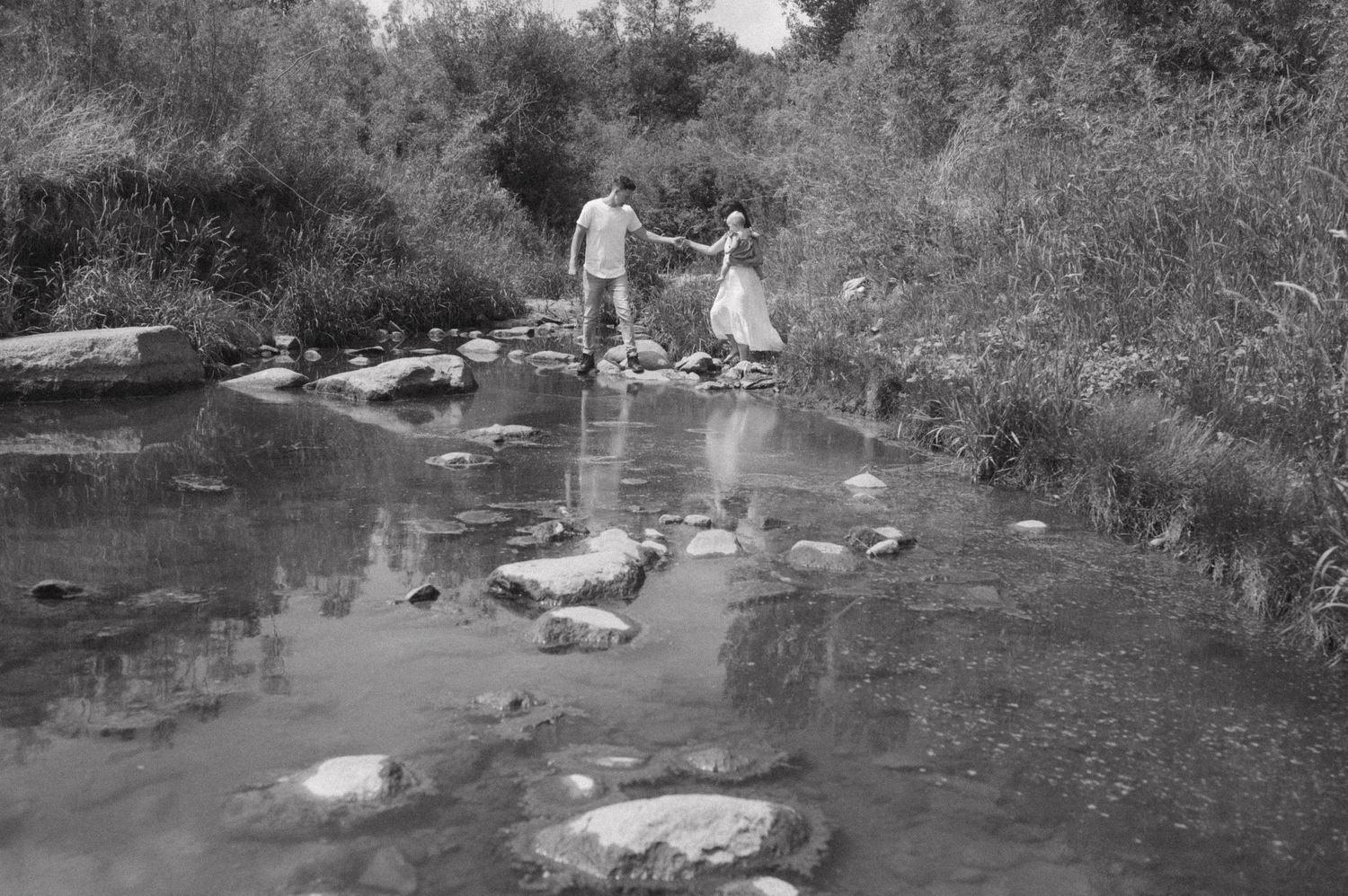 Black and white photo of couple crossing stream on stepping stones.