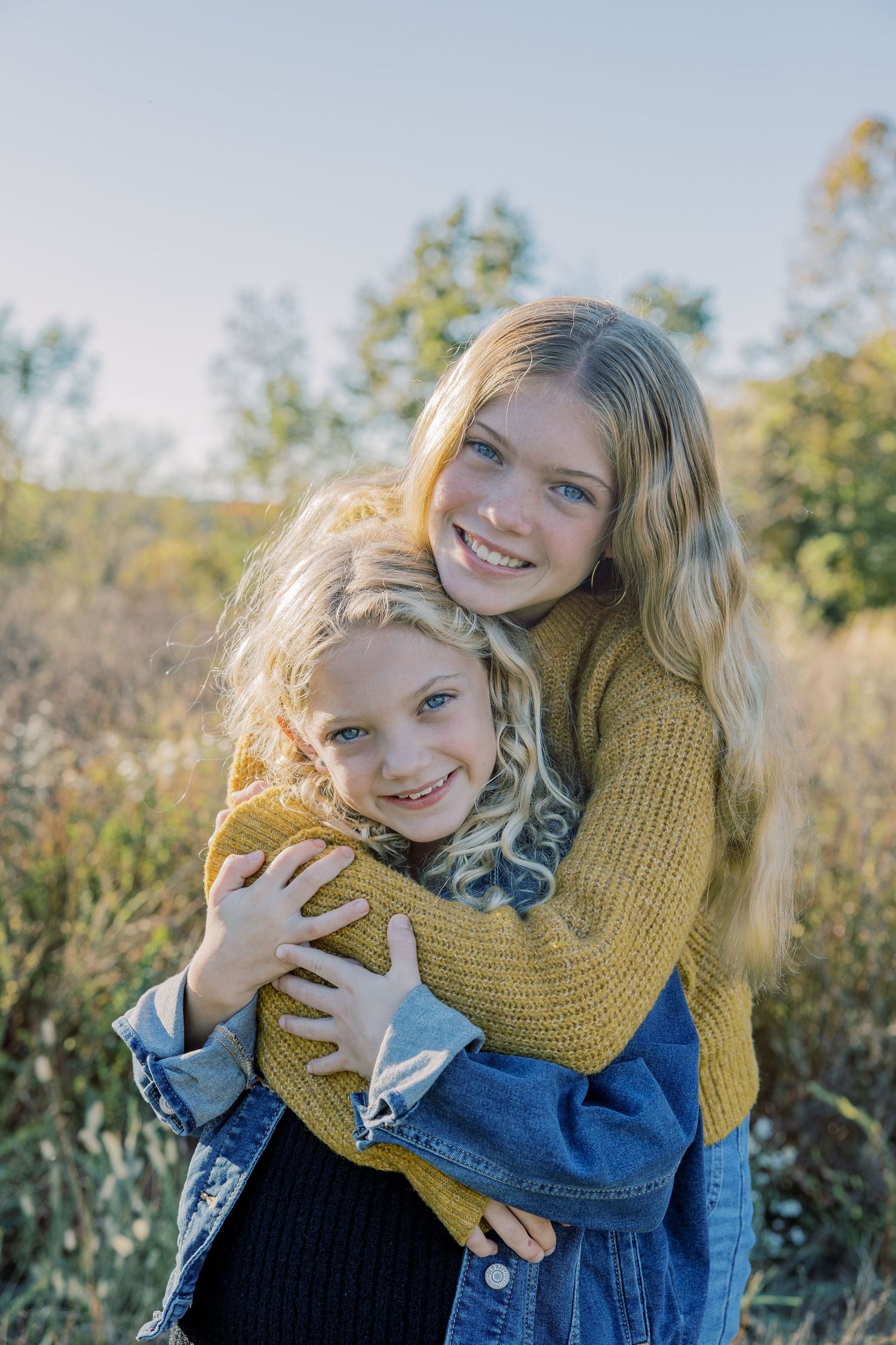 Two smiling blonde sisters hugging in an autumn field wearing cozy sweaters.