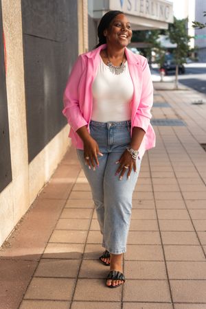 Person wearing pink blazer and jeans poses confidently on tiled walkway.