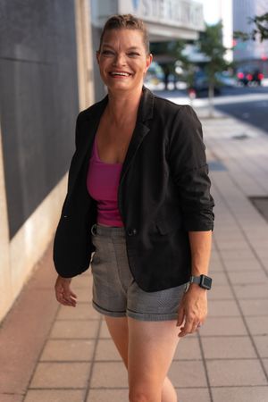Person wearing black blazer and pink top poses casually on urban sidewalk.